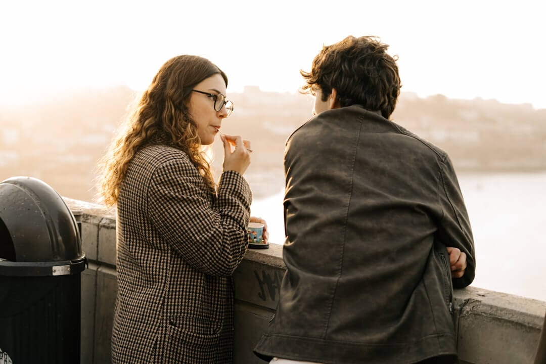 Two people sitting close on a balcony at dusk, talking gently under a soft evening sky